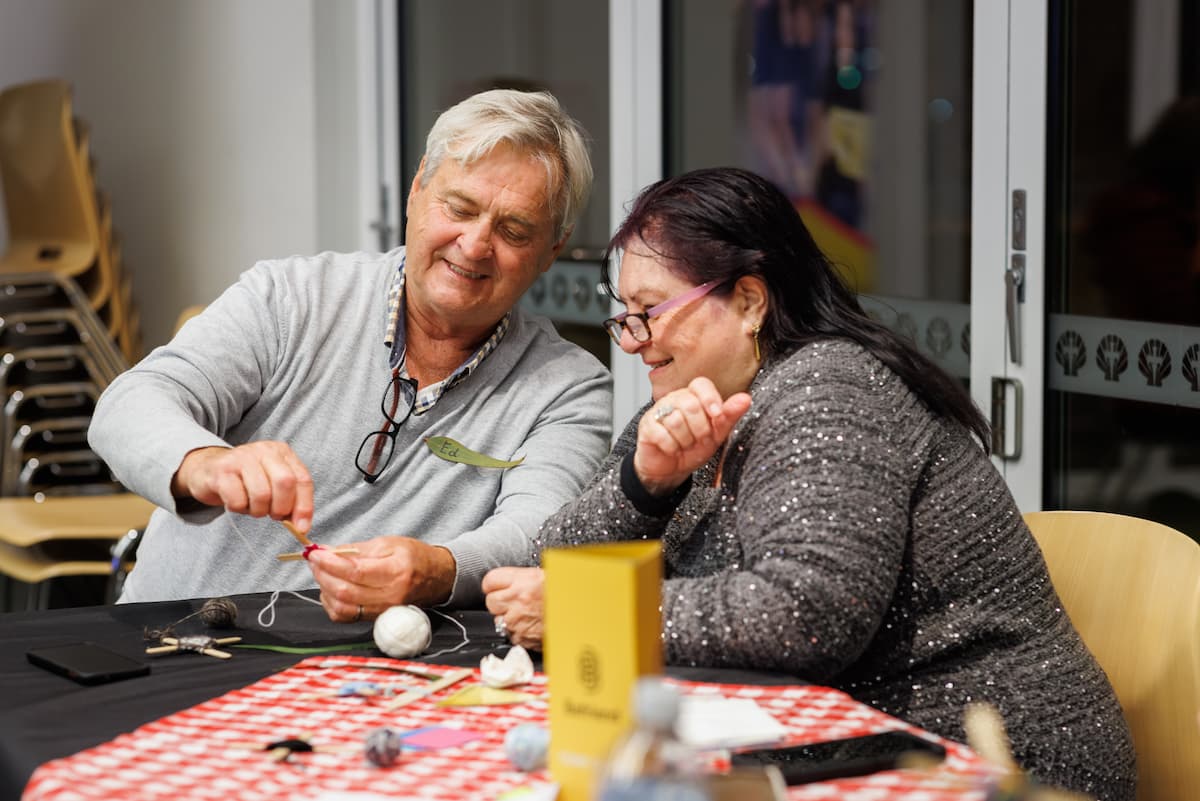 An man wearing a name tag reading "Ed" smiles as he ties string onto a small craft piece, while a woman beside him laughs, both seated at a table with yarn, ribbon and craft materials scattered across a red gingham cloth.